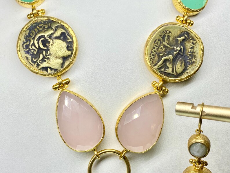 May include: Gold-toned necklace featuring coin-shaped pendants with embossed figures, paired with teardrop-shaped pink gemstones. The necklace includes a matching earring with a small, round, light-colored stone.