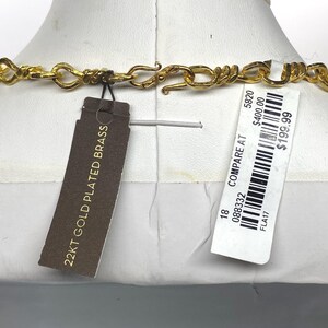 May include: A gold-colored necklace with a unique chain design is displayed on a mannequin. The necklace has a clasp closure. A tag reads "22KT GOLD PLATED BRASS". A price tag is also attached.