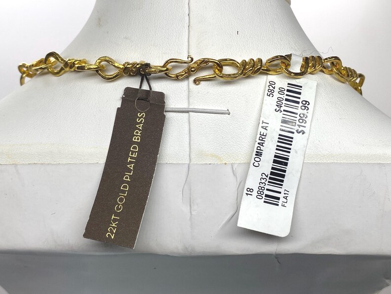 May include: A gold-colored necklace with a unique chain design is displayed on a mannequin. The necklace has a clasp closure. A tag reads "22KT GOLD PLATED BRASS". A price tag is also attached.