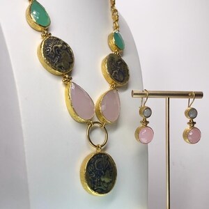 May include: A gold-toned necklace and matching earrings set. The necklace features green, pink, and coin-shaped pendants. The earrings have pink and gray stones. The jewelry is displayed on a white surface.