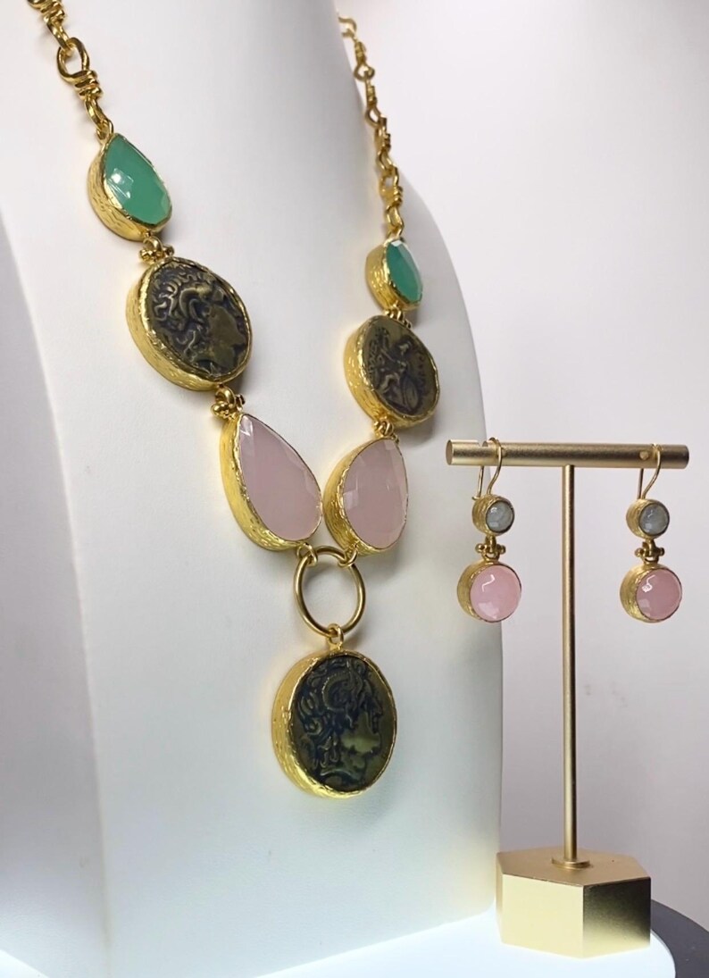 May include: A gold-toned necklace and matching earrings set. The necklace features green, pink, and coin-shaped pendants. The earrings have pink and gray stones. The jewelry is displayed on a white surface.