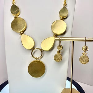 May include: A gold-toned necklace and matching earrings set. The necklace features a series of large, flat, circular and teardrop-shaped pendants. The earrings have a similar design, with a textured, circular pendant. The jewelry is displayed on a white bust and a gold stand.