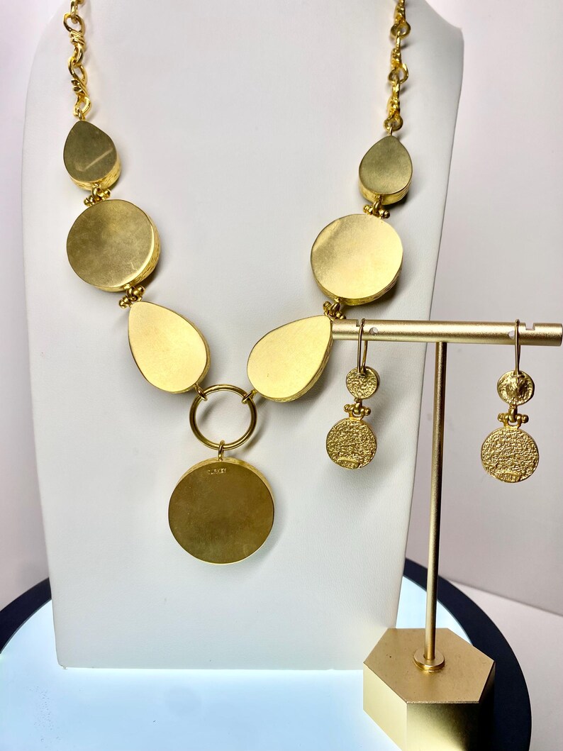 May include: A gold-toned necklace and matching earrings set. The necklace features a series of large, flat, circular and teardrop-shaped pendants. The earrings have a similar design, with a textured, circular pendant. The jewelry is displayed on a white bust and a gold stand.