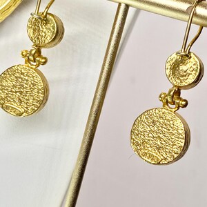 May include: Gold-toned dangle earrings with a textured, circular design. Each earring features a small, round top piece connected to a larger, textured disc below. The earrings are displayed on a gold-colored stand, showcasing their intricate details and craftsmanship.