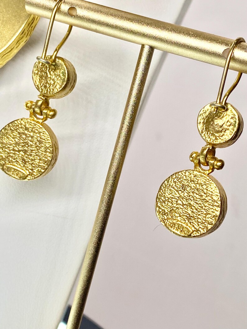 May include: Gold-toned dangle earrings with a textured, circular design. Each earring features a small, round top piece connected to a larger, textured disc below. The earrings are displayed on a gold-colored stand, showcasing their intricate details and craftsmanship.