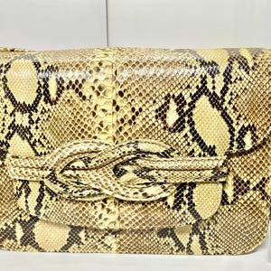 May include: A rectangular handbag with a snakeskin pattern in shades of tan, brown, and black. The bag features a knotted closure and a flap. The bag measures approximately 10 inches in length.