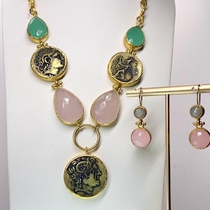 May include: A gold-toned necklace and matching earrings set. The necklace features green teardrop-shaped stones, coin-shaped pendants, and pink teardrop-shaped stones. The earrings have pink and gray stones. The jewelry is displayed on a white background.