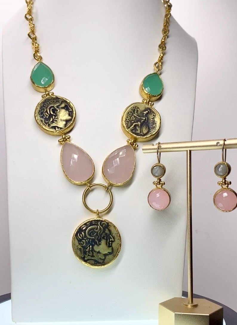 May include: A gold-toned necklace and matching earrings set. The necklace features green teardrop-shaped stones, coin-shaped pendants, and pink teardrop-shaped stones. The earrings have pink and gray stones. The jewelry is displayed on a white background.