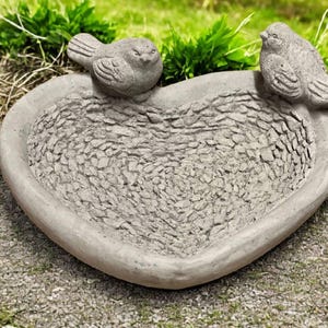 May include: A heart-shaped bird bath made of gray concrete. Two small bird sculptures are perched on the rim. The interior has a textured, stone-like appearance. The bird bath is designed for outdoor use.