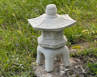 Può includere: Una lanterna di pagode in pietra grigia con un tetto decorativo e una base a quattro gambe. La lanterna è appoggiata su una roccia in un giardino.