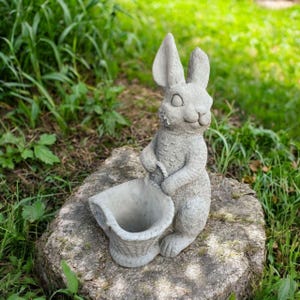 May include: A gray concrete rabbit statue holding a basket. The rabbit has long ears and is standing on a tree stump. The statue is outdoors, surrounded by green grass and foliage.