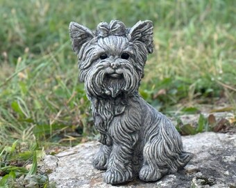 May include: A detailed, gray stone statue of a Yorkshire Terrier dog, sitting upright with its ears perked. The statue has a textured surface and is placed outdoors on a rock, with a blurred green background.