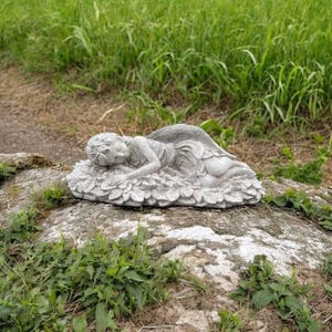 May include: A gray stone garden statue of a sleeping angel with detailed wings and a floral base. The angel is lying down, appearing peaceful. The statue is placed on a large rock, surrounded by greenery.