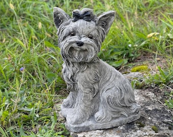 May include: A gray stone sculpture of a Yorkshire Terrier dog wearing a black bow. The sculpture is sitting on a rock with green grass in the background.