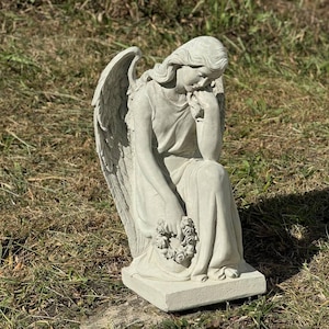 May include: A gray concrete statue of an angel with wings, sitting with a wreath of flowers in her hands. The angel is looking down with a sorrowful expression.