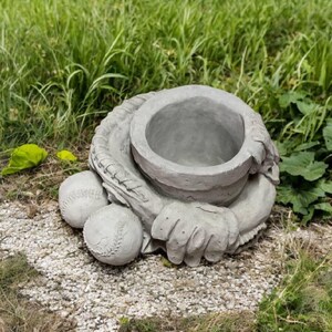 May include: A gray concrete baseball glove planter with two baseballs. The planter has a large opening for plants and is surrounded by green grass and small pebbles.