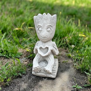 May include: A light gray, textured, anthropomorphic tree-like figure sculpture with large, round eyes and a smooth, smiling face. The figure is seated on a small, rectangular base and holds its hands together to form a heart shape at its chest.