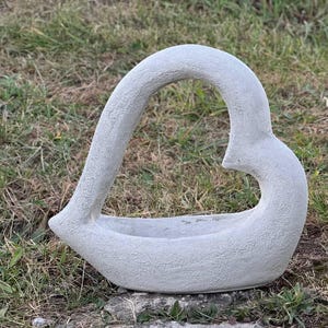 May include: A gray concrete heart-shaped planter with a smooth, textured surface. The planter is designed to hold plants or flowers.