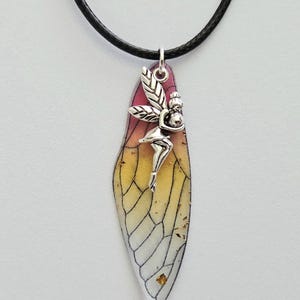 May include: A necklace featuring a black cord and a pendant shaped like an insect wing. The wing has a gradient of pink, orange, and yellow with black veins. A silver-toned metal fairy charm is attached to the top of the wing pendant.