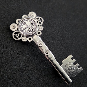 May include: A silver-toned metal key pendant featuring a circular clock face at the top, surrounded by an arrangement of various small mechanical gears. The shaft of the key is decorated with two small circular gear accents, and the bit has a gear design.