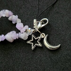 May include: A phone charm featuring a strand of irregular, light purple amethyst chip beads. Attached to the beads is a silver-toned metal lobster clasp holding a hollow star charm and a solid crescent moon charm, connected by a thin black cord.