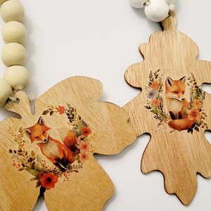 May include: Two wooden leaf-shaped ornaments with a fox and floral design. The ornaments are attached to a string of wooden beads. The fox is orange and white, surrounded by a wreath of flowers. The beads are light beige and white.