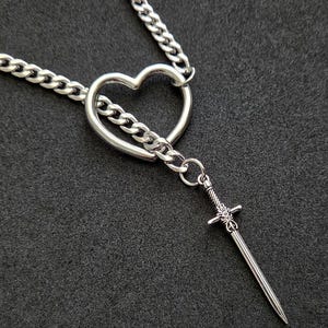 May include: A silver-toned metal necklace featuring a curb chain, a heart-shaped pendant, and a small sword charm. The chain passes through the heart, creating a lariat-style drop that suspends the sword pendant at the bottom of the assembly.