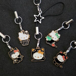 May include: Five metal phone charms featuring Hello Kitty characters in various Halloween costumes, including a skeleton, a vampire, a black cat, and a monster. Each charm is attached to a black cord with a silver-toned lobster clasp and ring.