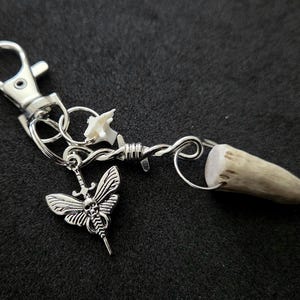 May include: A silver-toned keychain featuring a lobster clasp, a small white bone fragment, a silver charm shaped like a moth with a skull and sword motif, and a piece of textured, light brown antler attached to a twisted silver wire segment.