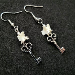 May include: Silver-toned key earrings with a decorative design. Each earring features a small, white, organic-shaped element above the key. The earrings hang from a simple hook.