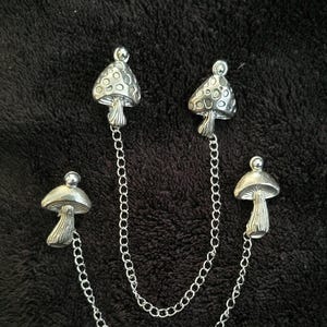 May include: A silver-colored mushroom brooch set with four mushroom charms connected by delicate chains. The mushroom caps have a textured design, and the brooch is set against a dark, textured background.