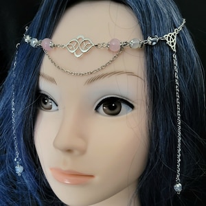 May include: A silver-toned headpiece with pink and clear beads, and a decorative Celtic knot design. The headpiece features a delicate chain that drapes down the side. The headpiece is displayed on a mannequin with blue hair.