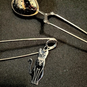 May include: A stainless steel heart-shaped tea infuser with a squeeze handle, and a silver cat charm pendant. The cat charm is black with silver details. The items are on a black background.