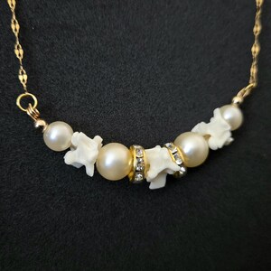 May include: A gold chain necklace featuring a central arrangement of white pearls, small white bone-shaped beads, and two gold-toned spacer beads encrusted with clear crystals. The necklace is displayed against a dark, textured background.