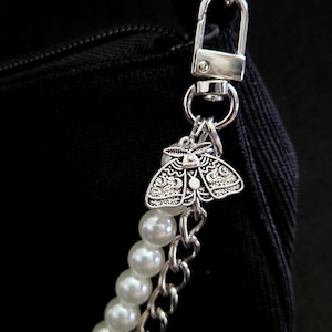 May include: A silver-toned metal moth charm with intricate wing patterns attached to a silver lobster clasp. The charm is connected to a strand of white faux pearls and a silver-toned metal chain, both secured to a black fabric bag.