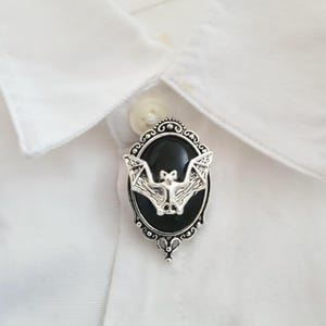 May include: A silver-toned brooch featuring an oval black cabochon set within an ornate, filigree-style frame. A detailed silver bat with outstretched wings is mounted centrally over the black stone, attached to a white collared shirt.