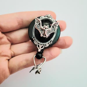 May include: A black and silver retractable badge reel with a bat design. The bat is silver and centered on a black circular background. The reel has a decorative silver frame and a black bead. A silver clip is attached.
