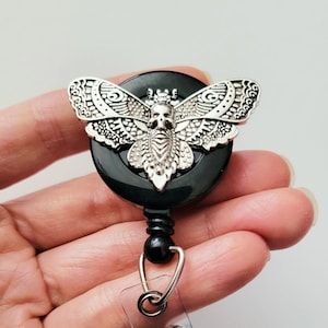 May include: A black circular badge reel featuring a silver-toned metal moth emblem. The moth has intricate wing patterns and a skull design on its thorax. A clear plastic strap with a metal snap button extends from the bottom of the reel.