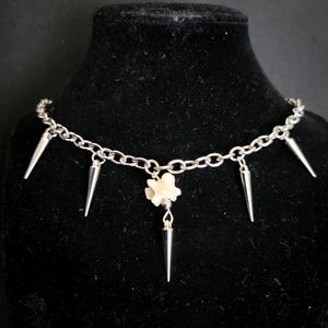 May include: Silver-toned choker necklace with a chain design. The necklace features five silver spikes and a small white flower pendant. The necklace is displayed on a black velvet surface.