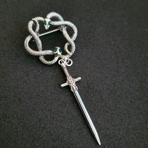 May include: Silver-toned brooch featuring a Celtic knot design with two snake heads and a dangling sword pendant. The snakes have green gemstone eyes. The brooch is set against a dark background.
