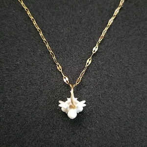 May include: A delicate gold-colored necklace with a small, white flower pendant. The pendant has a cluster of small, petal-like shapes around a central pearl-like bead. The chain has a delicate, linked design, and the background is a dark, solid color.