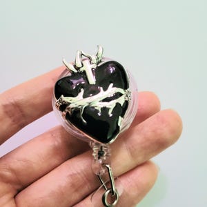 May include: A black heart-shaped badge reel with a silver cross and crown of thorns design. The badge reel has a clear plastic casing and a metal clip for attaching to clothing or accessories.