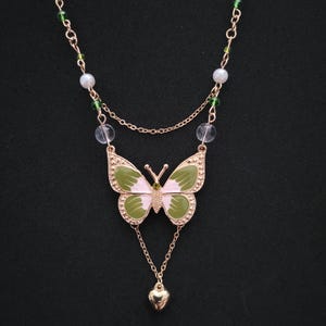 May include: A gold-toned necklace featuring a butterfly pendant with pink and green enamel wings. The necklace has a delicate chain with clear and green beads, and a small heart charm. The butterfly pendant is adorned with small, clear crystals.