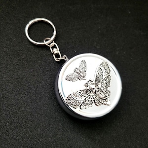 May include: A silver-colored keychain with a round, metallic charm featuring two detailed moth designs. The larger moth has a skull on its head. The keychain includes a metal ring for attaching keys.