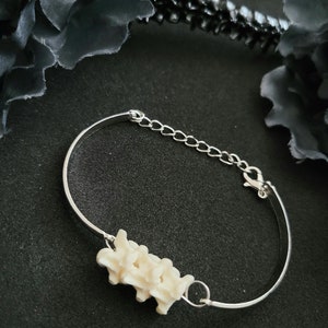 May include: A silver bracelet with a cream-coloured, spine-shaped charm. The bracelet includes an adjustable chain and a lobster clasp. The charm is detailed, offering a unique, gothic aesthetic. The bracelet is a distinctive piece of jewellery.