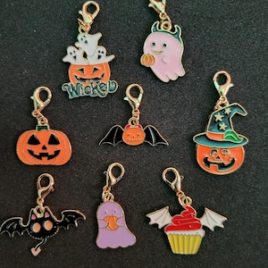 May include: A collection of Halloween-themed charms with gold-colored clasps. The charms feature various designs, including pumpkins, ghosts, bats, and a cupcake with bat wings. Some charms have the word "Wicked" on them. The charms are in orange, black, purple, and white.