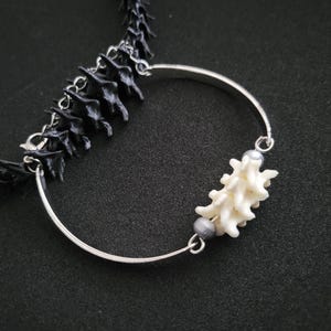 May include: A distinctive necklace with a black, spine-shaped chain and a silver-coloured, curved pendant. The pendant is decorated with a white, coral-like element and two grey beads. The necklace is set against a dark background.