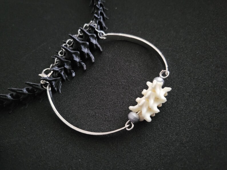 May include: A distinctive necklace with a black, spine-shaped chain and a silver-coloured, curved pendant. The pendant is decorated with a white, coral-like element and two grey beads. The necklace is set against a dark background.