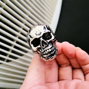 May include: A silver skull ring with detailed facial features, including eye sockets, a nose cavity, and teeth. The ring is held in a hand, showcasing its size and design. The skull ring is a gothic-inspired accessory.