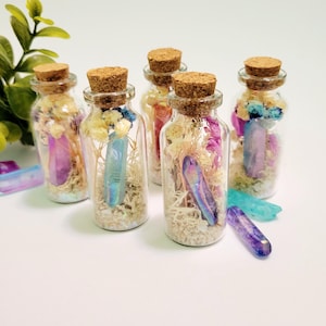 May include: Six small glass bottles with cork stoppers, each filled with dried flowers, moss, and a colorful crystal. The crystals are various colors, including purple, blue, and iridescent. The bottles are arranged on a white surface.